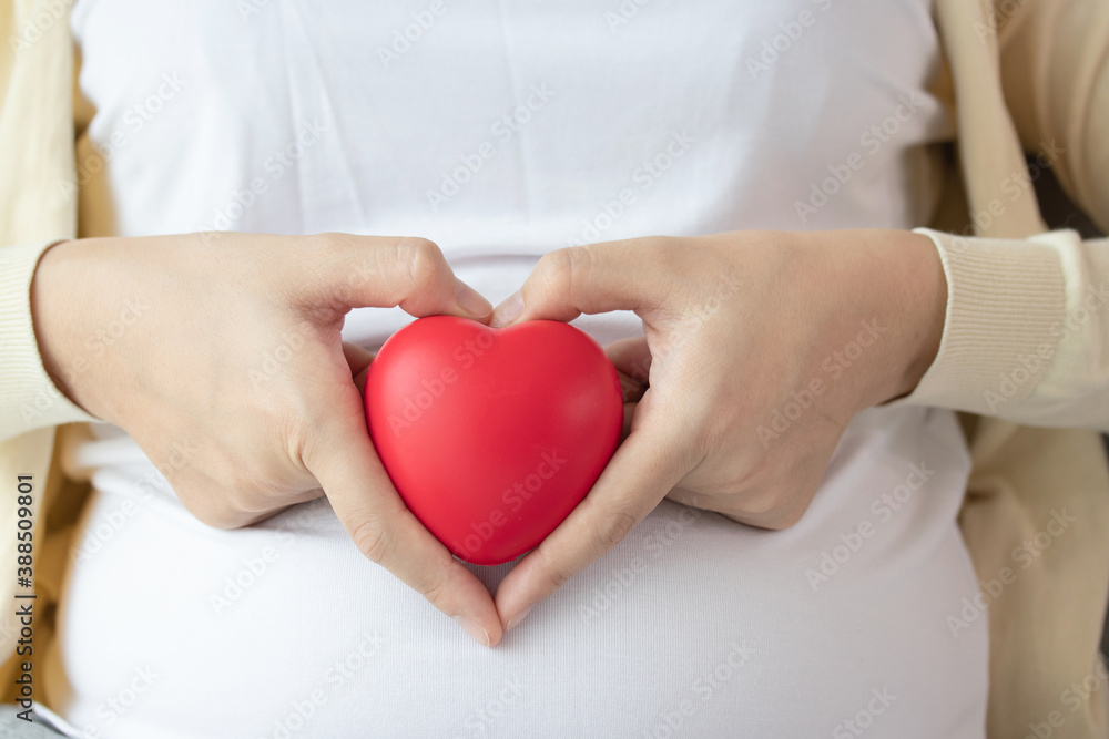 Beautiful pregnant woman sitting on bed and making hand heart on red heart ball at home. Pregnancy, motherhood, love and expectation concept. Beautiful pregnant woman sitting on bed and making hand heart on red heart ball at home. Pregnancy, motherhood, love and expectation concept.
