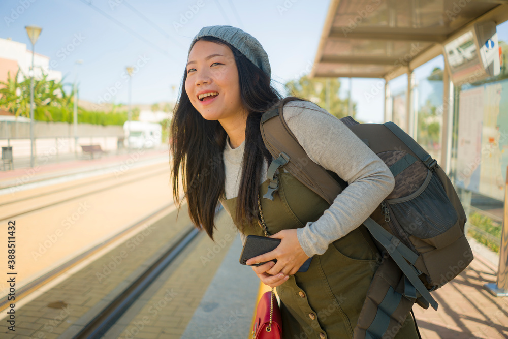 Fototapeta premium outdoors portrait of young happy and beautiful Asian Chinese tourist woman with backpack holding mobile phone on train station waiting excited in holidays travel and vacation