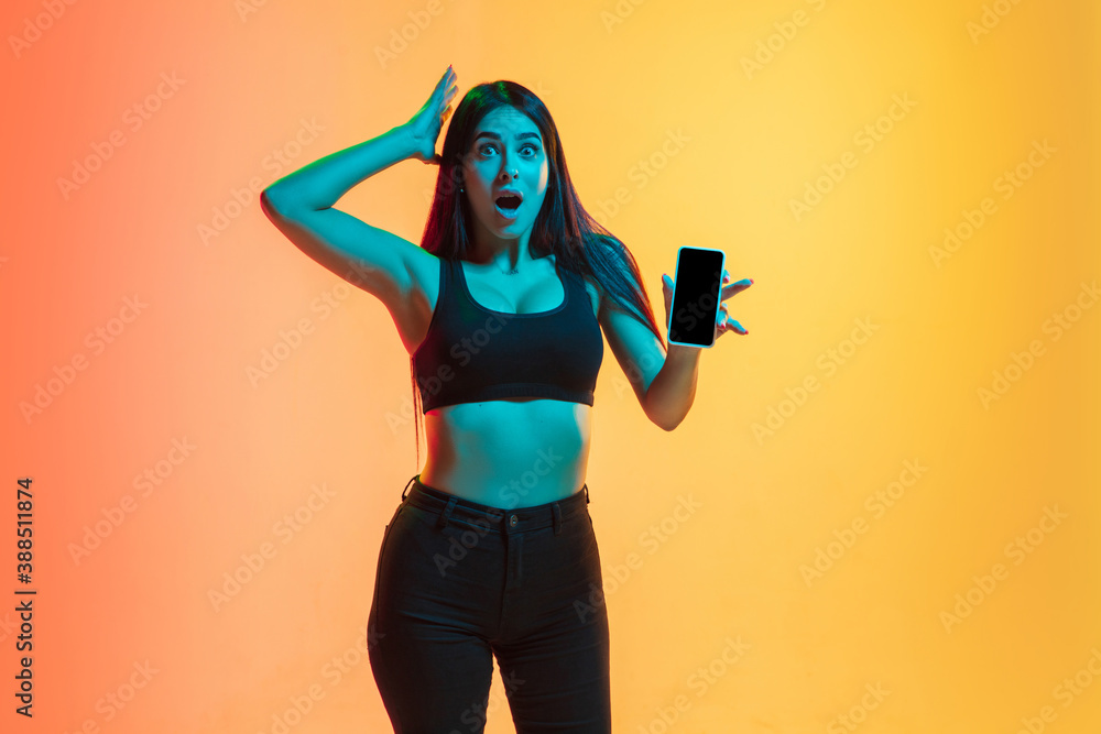 Obraz premium Showing phone's screen. Young caucasian woman's portrait on gradient yellow-orange studio background in neon light. Concept of youth, human emotions, facial expression, sales, ad. Sportive fit model.