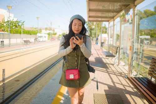 outdoors portrait of young happy and beautiful Asian Chinese tourist woman with backpack using mobile phone on train station waiting excited in holidays travel and vacation