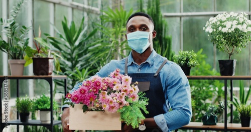 Close up portrait of African American happy handsome man owner of flower shop in face mask smiling and holding floral box in hands. Young joyful man manager in greenhouse with olants. Worker concept photo