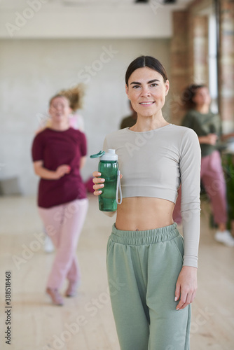 Portrait of young female athlete in sports clothing drinking water and smiling at camera standing in dance studio