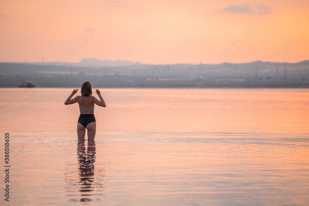 A girl in a bikini stands in the water at sunset. Rear view.