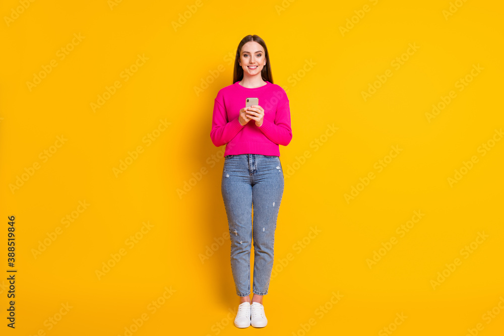 Full length photo of charming lady hold telephone look camera beaming smile wear pink sweater jeans sneakers isolated yellow color background Full length photo of charming lady hold telephone look camera beaming smile wear pink sweater jeans sneakers isolated yellow color background