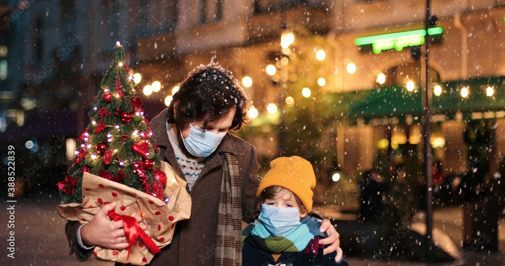 Portrait of Caucasian happy man hugging little son while walking outdoor. Joyful parent with small boy on street in snow town with little new year tree and present in good mood. Christmas concept