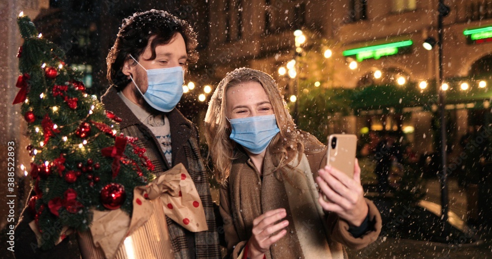 Portrait of Caucasian joyful wife and husband in masks making selfie photo while snowing on street. Happy family talking on video on smartphone with new year tree outdoor?. Holidays. Christmas concept