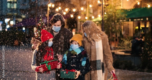 Portrait of Caucasian happy family walking with presents outdoors. Joyful kids with parents on street in city with little new year tree while snowing. Holiday lights and decoration. Christmas concept