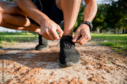 Male athlete bending down tying exercise shoe laces before training with smartwatch to track fitness outdoors on running path in sun