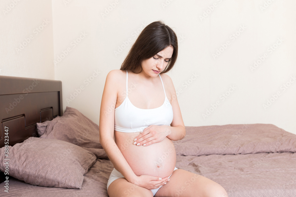 Young pretty pregnant woman, in pain, sitting on bed in bedroom, preparing to give a birth Young pretty pregnant woman, in pain, sitting on bed in bedroom, preparing to give a birth