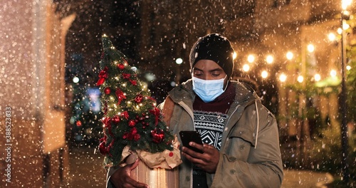 Close up portrait of African American handsome male in mask texting on smartphone while standing on crowded street. Man with christmas tree tapping on cellphone while snowing. New year concept