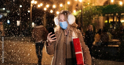 Close up portrait of Caucasian young beautiful female in medical mask texting on smartphone while standing on street. Happy woman with christmas gift tapping on cellphone while snowing © VAKSMANV
