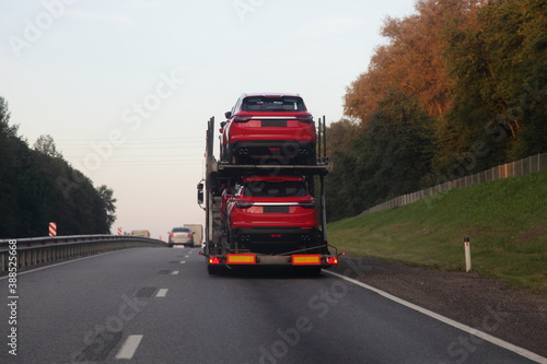 Loaded two level car carrier truck with car transporter semi trailer drive on suburban highway road, rear view close up, delivery new red chinese autos logistics, automobile transportation