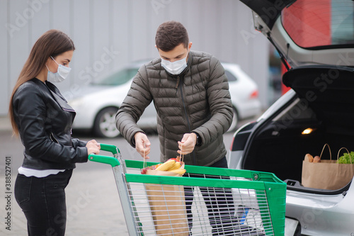 Young stylish couple in protectivemask with shopping cart full of fresh food, packing products into the car on the outdoor parking. Shopping during quarantine photo
