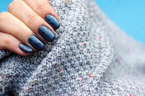 Female hand in a gray knitted sweater fabric with beautiful manicure - dark gray blue glittered nails with copy space. Selective focus. Closeup view