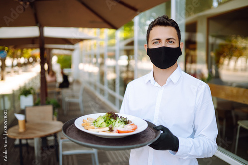 A young waiter in a protective mask on his face and gloves holding plate with food in cafe. Waiter serving in motion on duty in restaurant.  Covid- 2019.