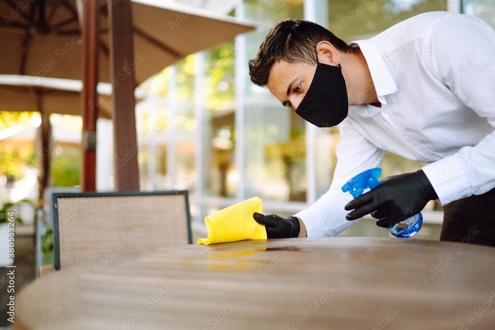 Waiter in protective face mask and gloves cleaning the table with disinfectant spray in a restaurant. Covid- 2019. Waiter in protective face mask and gloves cleaning the table with disinfectant spray in a restaurant. Covid- 2019.
