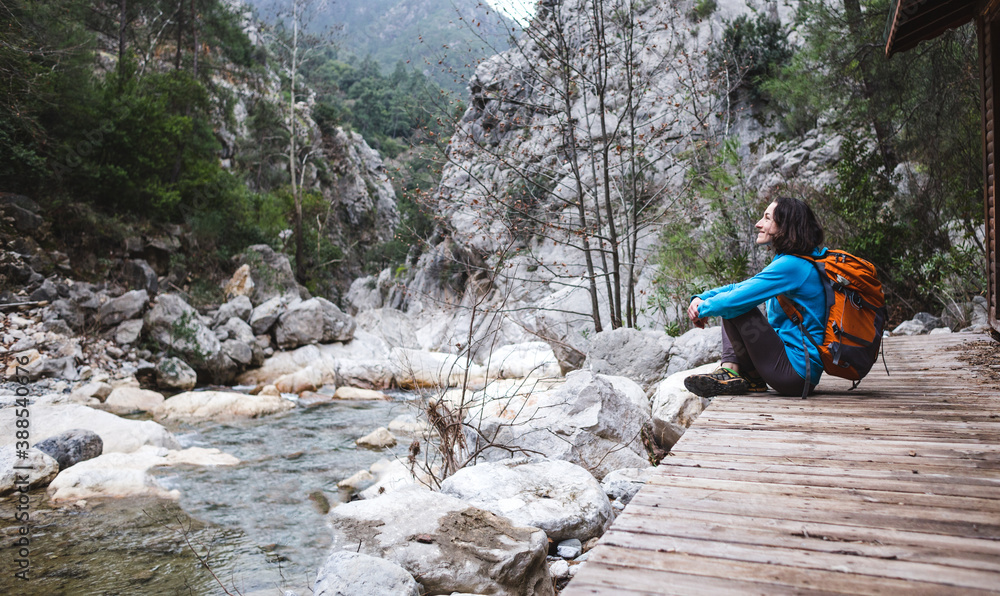 A woman with a backpack sits on the porch of an old wooden house and looks at a mountain river.