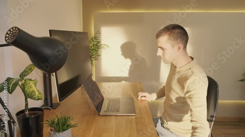 Panning shot of a young professional moving with the desk as he transitions from sitting to standing at an idyllic home working setup. Beautiful evening light on his face and plants fill the desk.