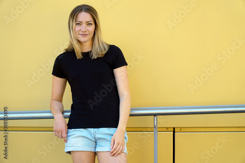 Leinwand Poster Attraktive blonde Frau vor gelber Wand trägt T-Shirt in schwarz mit Copy Space o