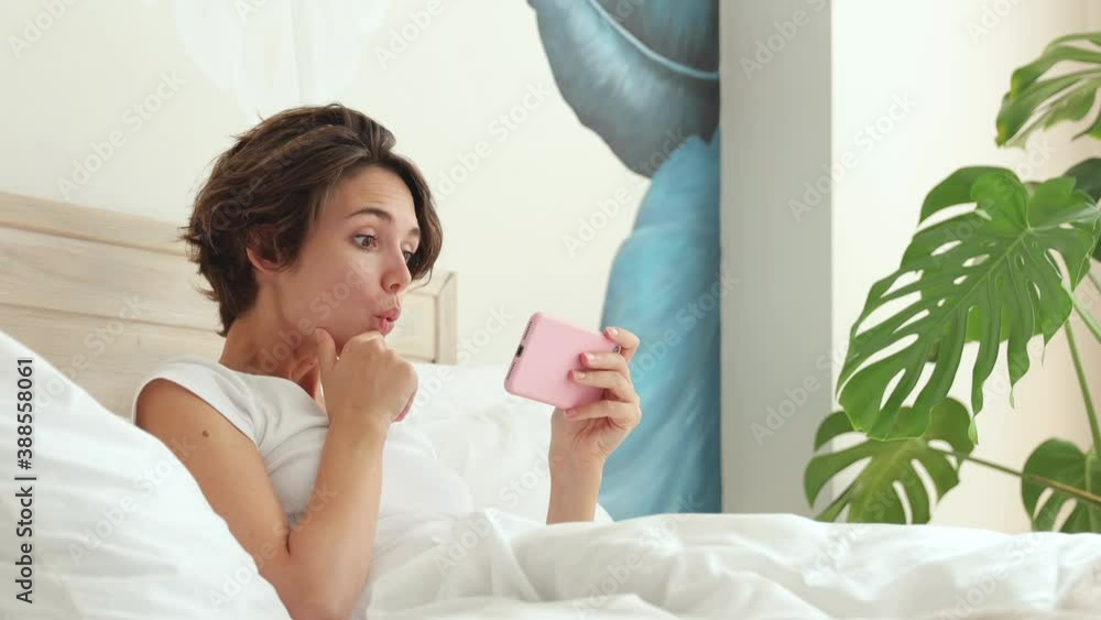 Young woman in white t-shirt pajamas home wear lying in bed spending time in bedroom holding using mobile cell phone get video call videoconference isolation quarantine covid-19 Rest lifestyle concept Young woman in white t-shirt pajamas home wear lying in bed spending time in bedroom holding using mobile cell phone get video call videoconference isolation quarantine covid-19 Rest lifestyle concept