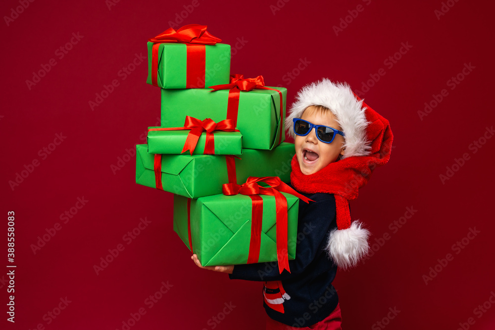 little boy in sunglasses and a Santa hat holds a pyramid of Christmas gift boxes little boy in sunglasses and a Santa hat holds a pyramid of Christmas gift boxes