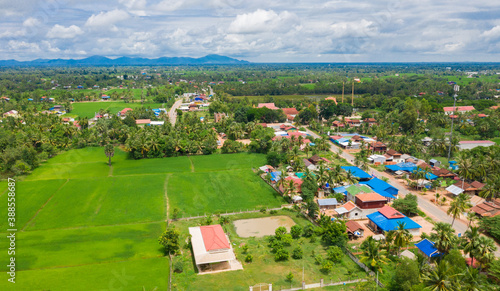 A top down aerial view of a small country town with traditional houses with orange roofs  a red dirt road  rice fields  and palm trees in the jungle in Cambodia.