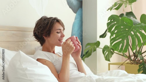 Calm young woman in white t-shirt pajamas home wear lying in bed spending time in bedroom hold cup of coffee drink wake up feel good better regain consciousness. Rest relax good mood lifestyle concept