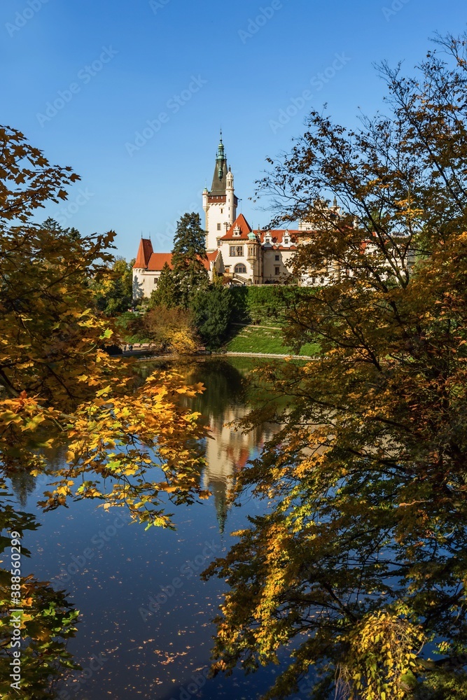 Pruhonice, Czech Republic - October 25 2020: Scenic view of romantic castle standing in a park surrounded with yellow and orange trees. Reflection of the castle in water. Sunny autumn day, blue sky.