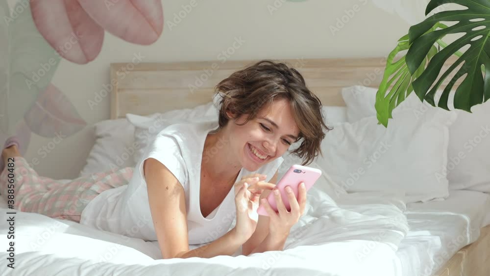 Dreamful calm young woman in white t-shirt pajamas home wear lying in bed spending time in bedroom hold in hand using mobile cell phone send typing sms message. Rest relax good mood lifestyle concept Dreamful calm young woman in white t-shirt pajamas home wear lying in bed spending time in bedroom hold in hand using mobile cell phone send typing sms message. Rest relax good mood lifestyle concept