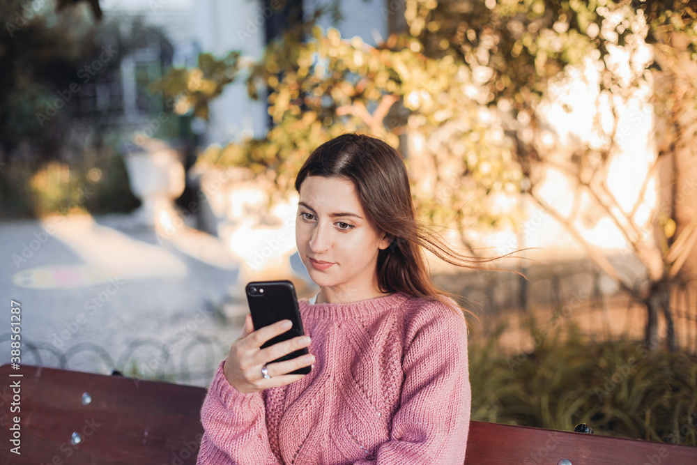 Young woman in pink sweater looking at her smartphone and sitting on a bench Young woman in pink sweater looking at her smartphone and sitting on a bench