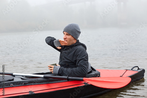 Handsome sitting in kayak, wakes up early in morning to rowing boat and doing water sport, guy yawning and hold mug with hot tea as have rest while canoeing, cover his mouth with his hand.
