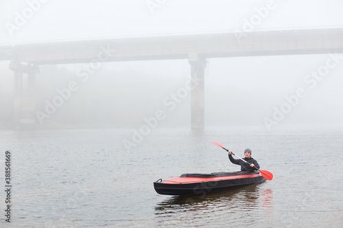 Man canoeing in traditional wooden kayak on large lake at cold cloudy day, fog over water, handsome guy wearing black jacket and gray cap, kayaking alone in lake with bridge on backround.
