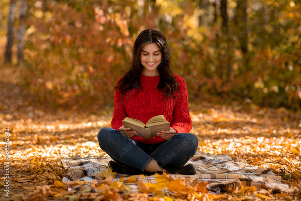 Charming young woman in knitted red sweater reading book while sitting on picnic blanket at autumn park Charming young woman in knitted red sweater reading book while sitting on picnic blanket at autumn park
