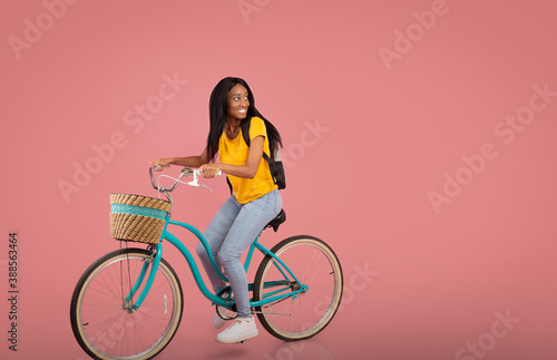 Pretty black lady riding bicycle and looking aside at empty space over pink studio background © Prostock-studio