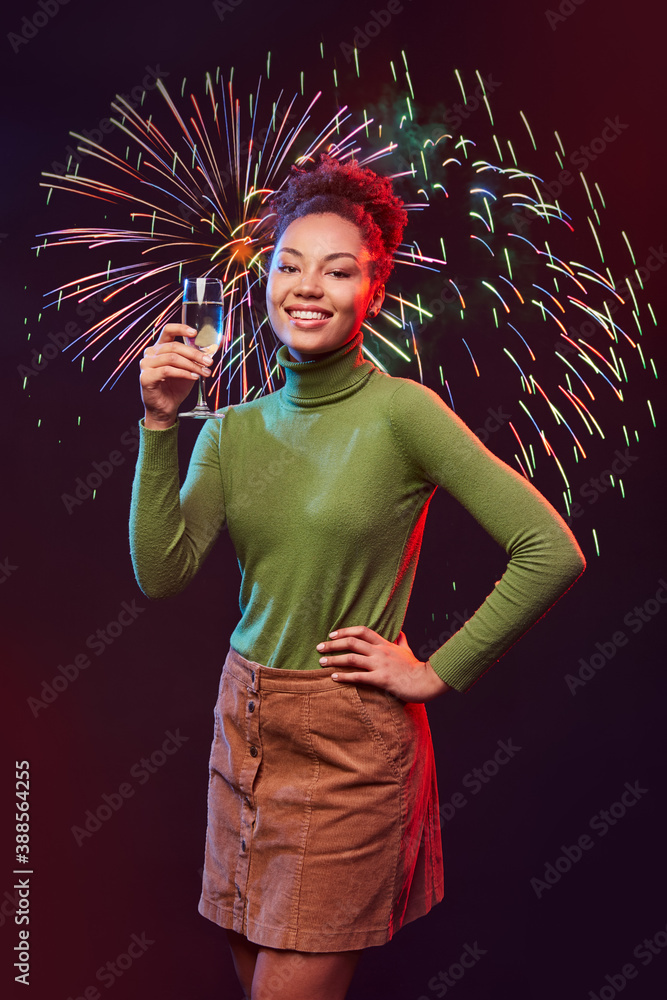 Fototapeta premium Vertical shot of a young happy mixed race woman with glass of champagne standing against fireworks background, smiling at camera