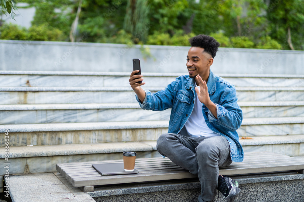 Smiling young african man in casual clothes making video call with smartphone and gesturing greeting while sitting on bench in park Smiling young african man in casual clothes making video call with smartphone and gesturing greeting while sitting on bench in park