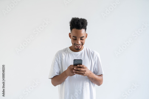 Portrait of young african american man isolated on gray background wearing white t-shirt standing in front of camera, looking attentively with smile at screen of smartphone he is holding