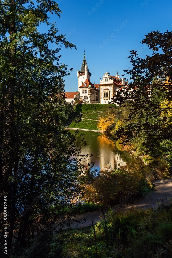 Pruhonice, Czech Republic - October 25 2020: View of the romantic castle surrounded with green and yellow trees standing in park. Reflection of the castle in water. Bright sunny autumn day, blue sky.