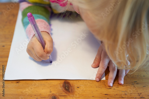 4 year old blonde girl with a brightly colored striped sweater sits at a wooden table and draws with a purple pencil on a piece of white paper