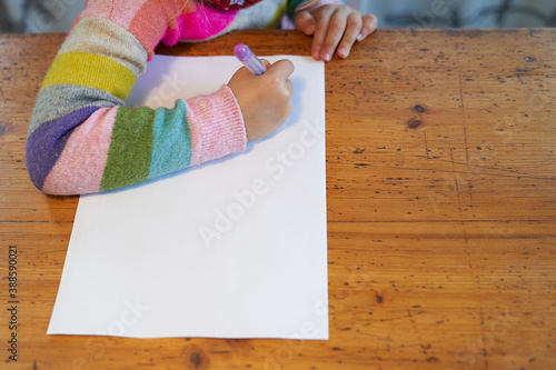 4 year old blonde girl with a brightly colored striped sweater sits at a wooden table and draws with a purple pencil on a piece of white paper