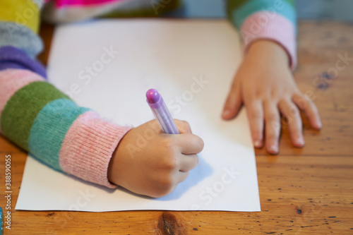 4 year old blonde girl with a brightly colored striped sweater sits at a wooden table and draws with a purple pencil on a piece of white paper