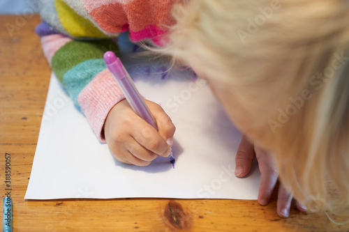 4 year old blonde girl with a brightly colored striped sweater sits at a wooden table and draws with a purple pencil on a piece of white paper