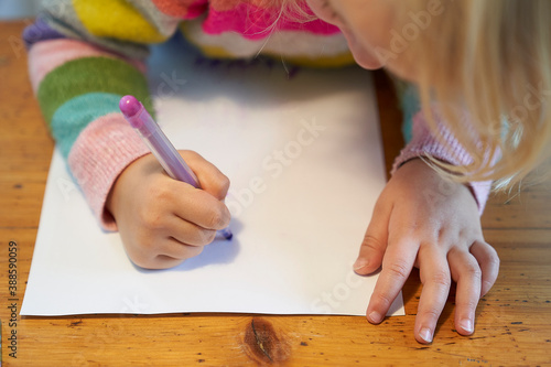 4 year old blonde girl with a brightly colored striped sweater sits at a wooden table and draws with a purple pencil on a piece of white paper
