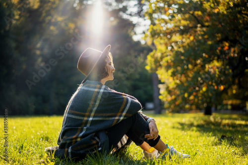 Portrait of a beautiful curly young woman in an autumn park in a hat and a plaid scarf sitting on the grass enjoying nature