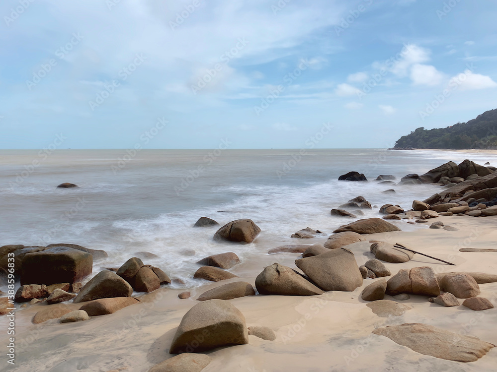 Slow shutter shot of waves crashing into rocks on a beach on a clear day. Small mountain in the background on the right with blue sky. Slow shutter shot of waves crashing into rocks on a beach on a clear day. Small mountain in the background on the right with blue sky.