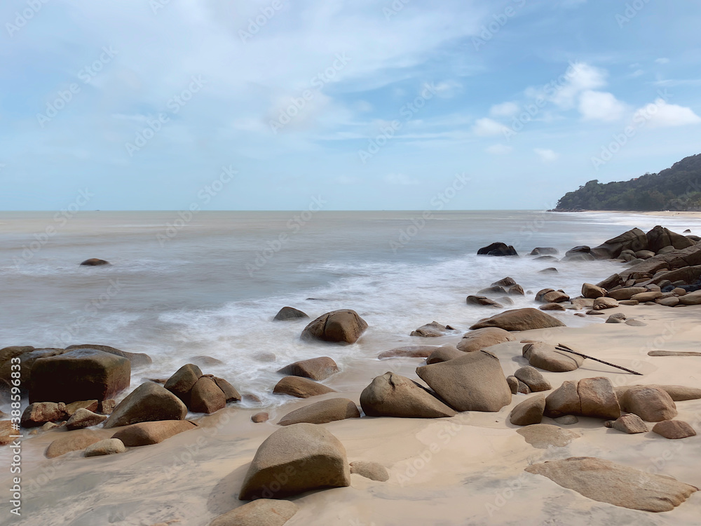 Slow shutter shot of waves crashing into rocks on a beach on a clear day. Small mountain in the background on the right with blue sky.