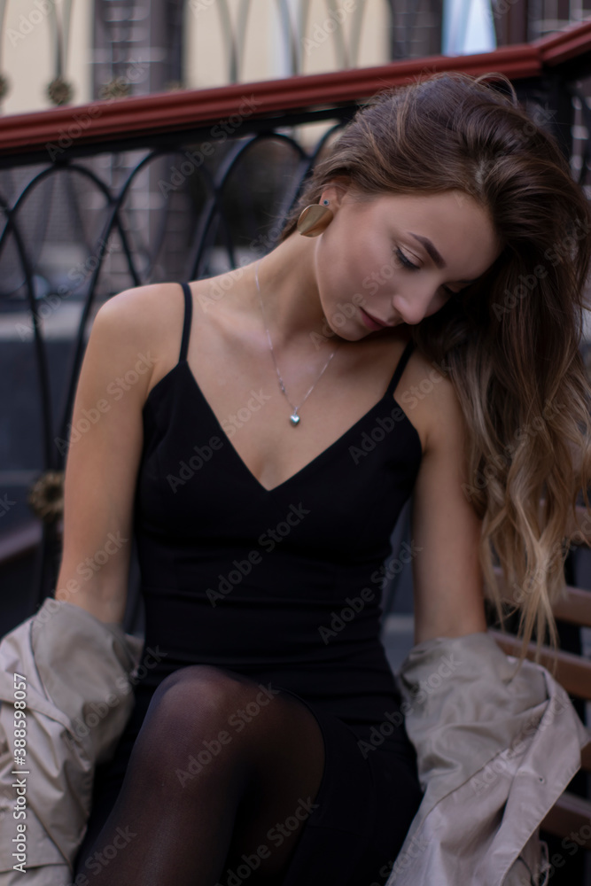 Fototapeta premium attractive slim young brunette in fashionable trench coat and black dress sitting on a bench. autumn, yellow leaves.