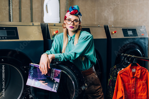 Fotografia Trendy woman in glasses and turban holding magazine in laundromat