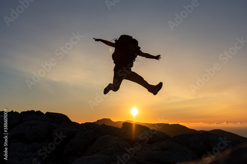 Horizontal image of hiker at the top of a mountain jumping on the sun of the beautiful golden hour sunset with intense color and blue sky fused with yellow, carrying large backpack