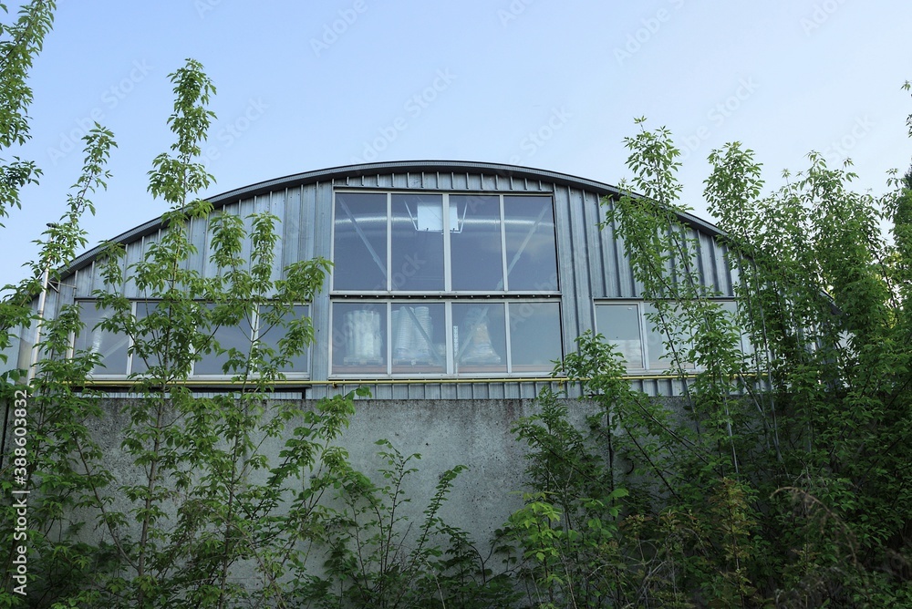 gray metal wall of a factory with a large window against the sky behind a concrete fence in green vegetation gray metal wall of a factory with a large window against the sky behind a concrete fence in green vegetation